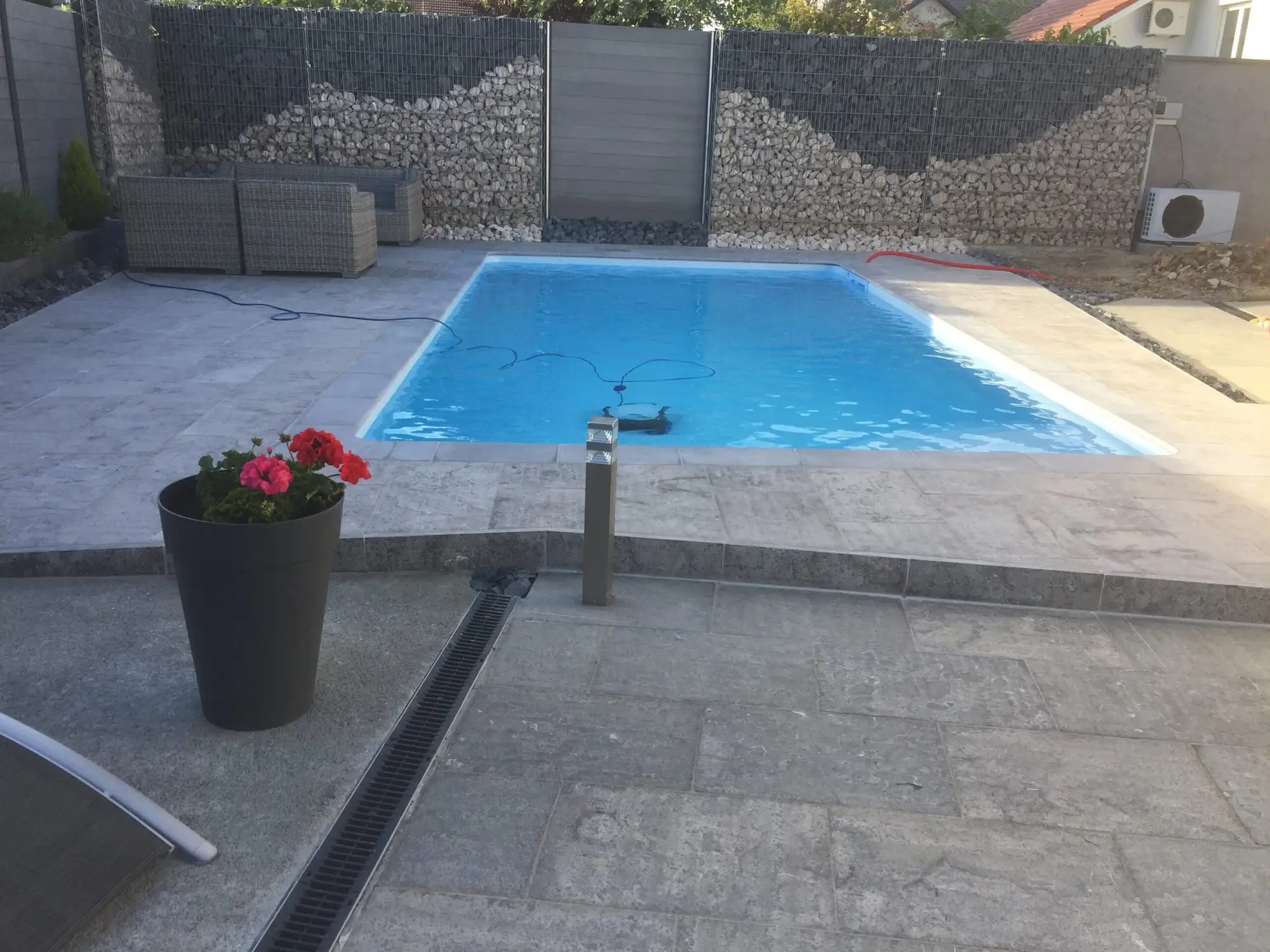 Plage de piscine en dallage gris avec caniveau, bordée d’un mur en gabions