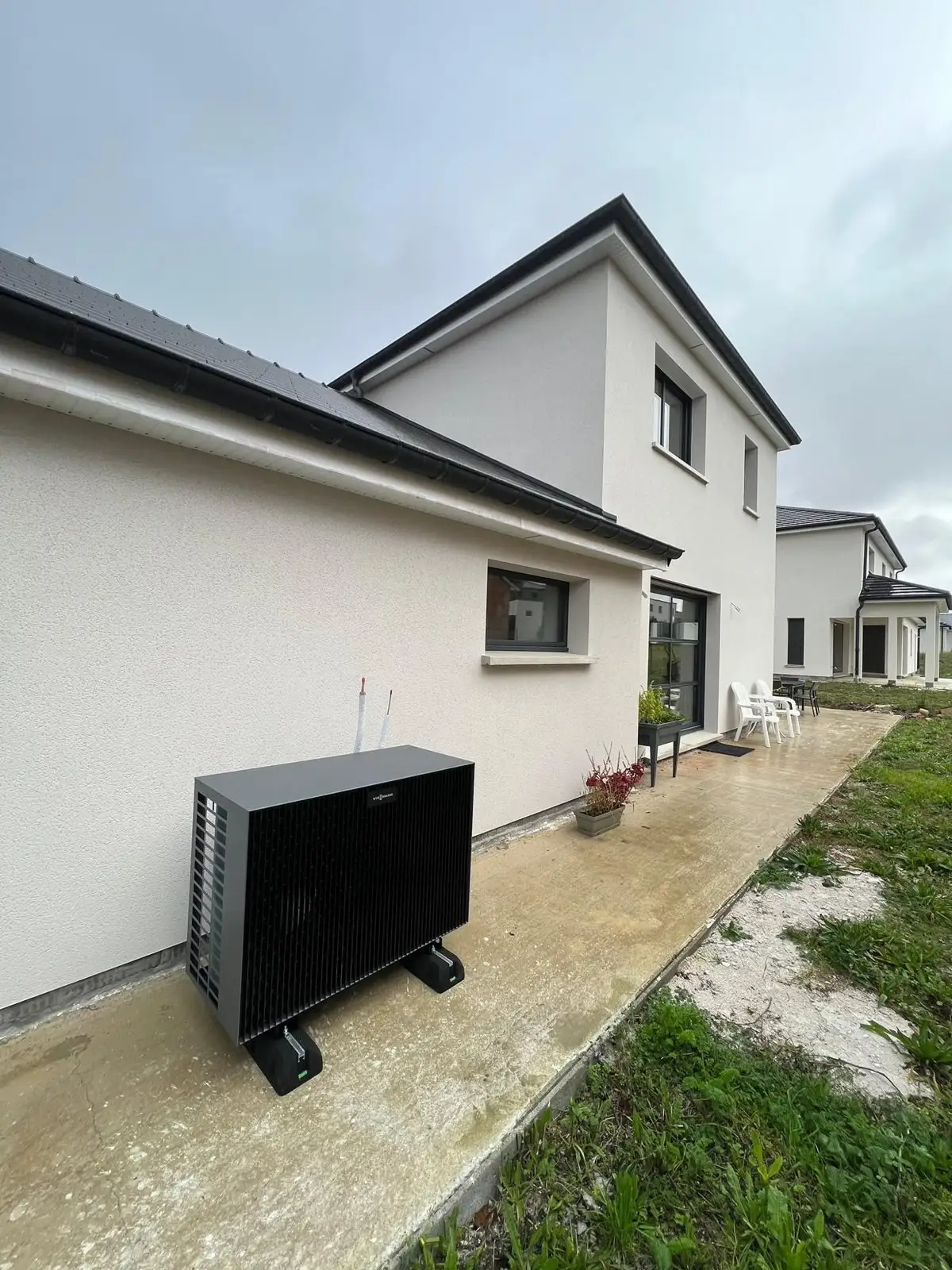 Unité extérieure de pompe à chaleur posée sur plots le long d’une maison récente enduite en blanc